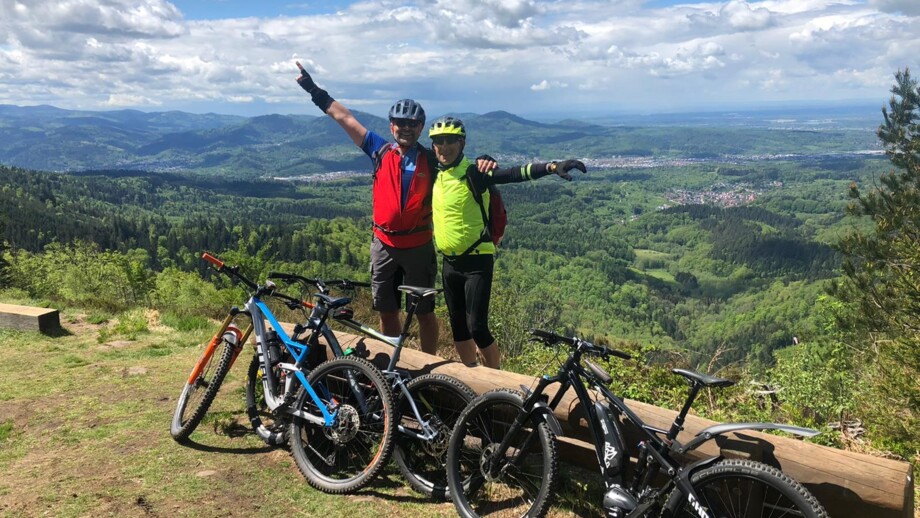 Zwei strahlende Mountainbiker nach Ankunft am Gipfel mit der Aussicht über den Schwarzwald bei sonnigem Wetter.