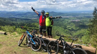 Zwei strahlende Mountainbiker nach Ankunft am Gipfel mit der Aussicht über den Schwarzwald bei sonnigem Wetter.