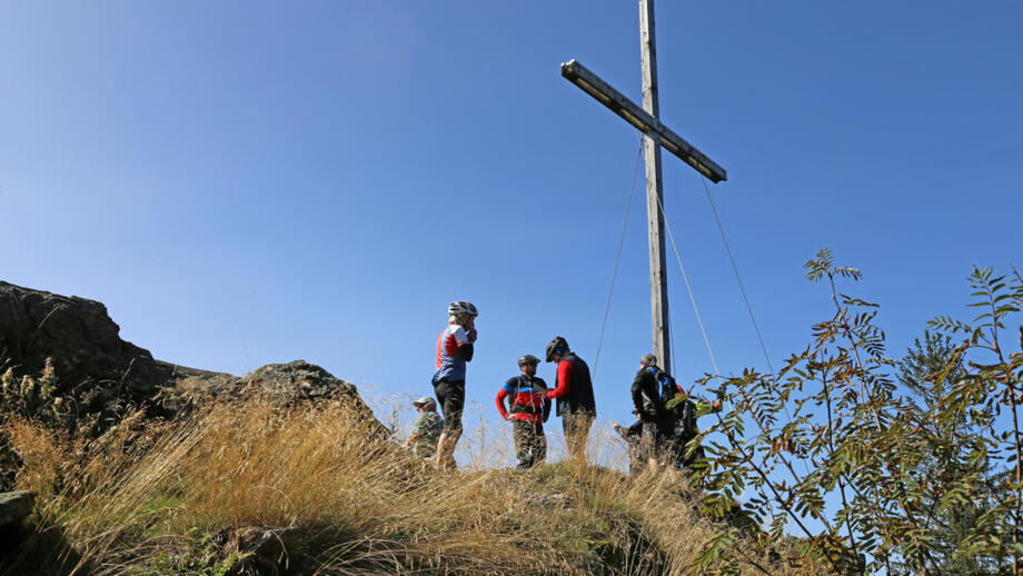 Ein Mountainbike Gruppe an einem Gipfelkreuz. Der Himmel ist blau und die Sonne scheint.