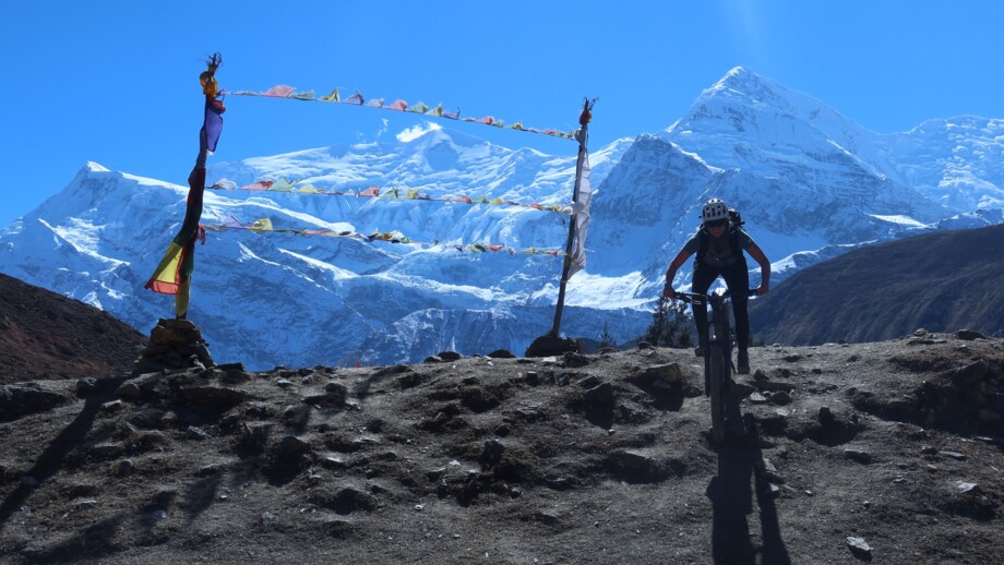 Ein Mountainbiker unterwegs auf der Annapurna-Umrundung in Nepal. In Hintergrund sieht man die 8000er des Himalayas. Gebetsfahnen wehen im Wind.