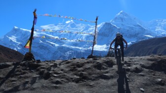 Ein Mountainbiker unterwegs auf der Annapurna-Umrundung in Nepal. In Hintergrund sieht man die 8000er des Himalayas. Gebetsfahnen wehen im Wind.