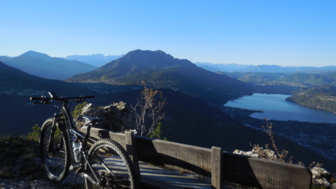 Ein Mountainbike an einen Zaun angelegt mit Ausblick auf die umliegende Bergwelt und den See.