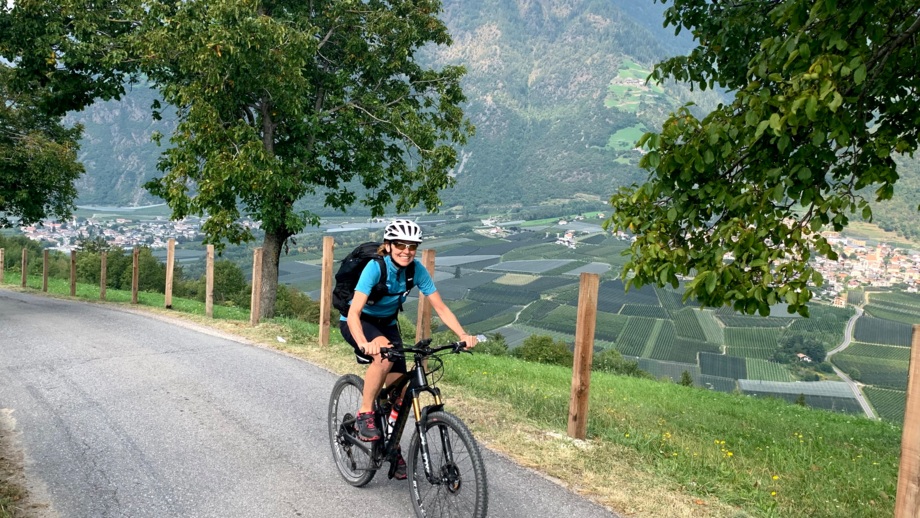 Eine Mountainbikerin fährt auf einer Asphaltstrasse. Im Hintergrund sind die Berge zu sehen.