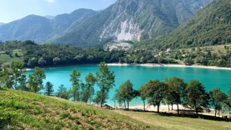 Ein türkisblauer See mit der Bergwelt im Hintergrund. Der Himmel ist leicht bewölkt mit Haufenwolken. Die Sonne scheint.