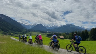 Eine Gruppe an Mountainbiker fährt auf einer Strasse. Am Wegesrand sind die grünen Allgäuer Wiesen zu sehen. Im Hintergrund die Berge.