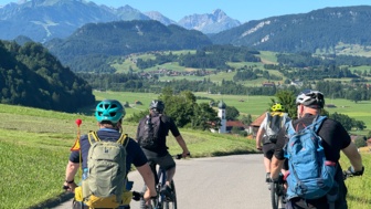 Eine Gruppe an Mountainbiker unterwegs auf Tour.  Sie fahren auf einer Straße. Die Sonne scheint. Im Hintergrund sieht man die Berge.