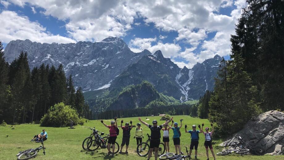 Eine Gruppe an Mountainbiker in Slowenien. Sie stehen auf einer Wiese. Im Hintergrund sind die Berge zusehen. Der Himmel ist bewölkt. Die Sonne scheint.