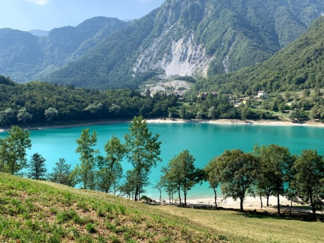 Ein See in den Bergen, das Wasser ist türkisblau. Im Hintergrund sind die Berge zu sehen. Die Sonne scheint.
