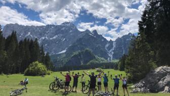 Eine MTB-Gruppe steht auf einer Wiese und hebt die Arme in die Höhe. Sie sind glücklich über Ihre Tour. Der Himmel ist bewölkt, die Sonne scheint. Im Hintergrund sieht man die Bergwelt des Triglav Nationalsparks.