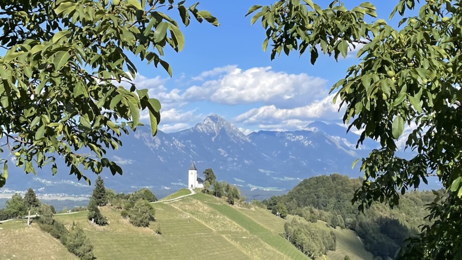 Eine Kirche auf einer Anhöhe stehend. Zur Kirche führt ein Pfad. Der Himmel ist blau. Die Sonne scheint. Am Himmel sind wenige Wolken. Im Hintergrund sind die Berge Sloweniens zu sehen.