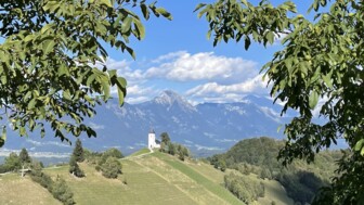 Eine Kirche auf einer Anhöhe stehend. Zur Kirche führt ein Pfad. Der Himmel ist blau. Die Sonne scheint. Am Himmel sind wenige Wolken. Im Hintergrund sind die Berge Sloweniens zu sehen.