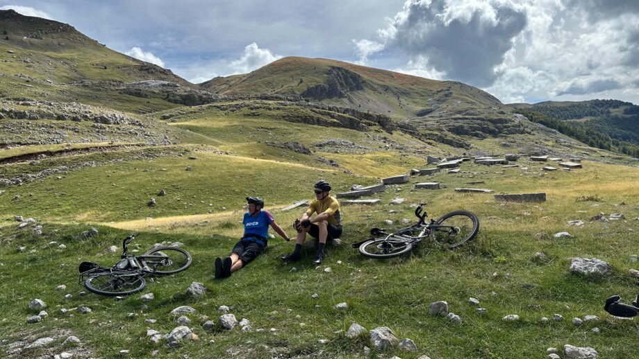 Zwei Mountainbiker machen eine Pause auf einer Wiese. Im Hintergrund sind die Berge.