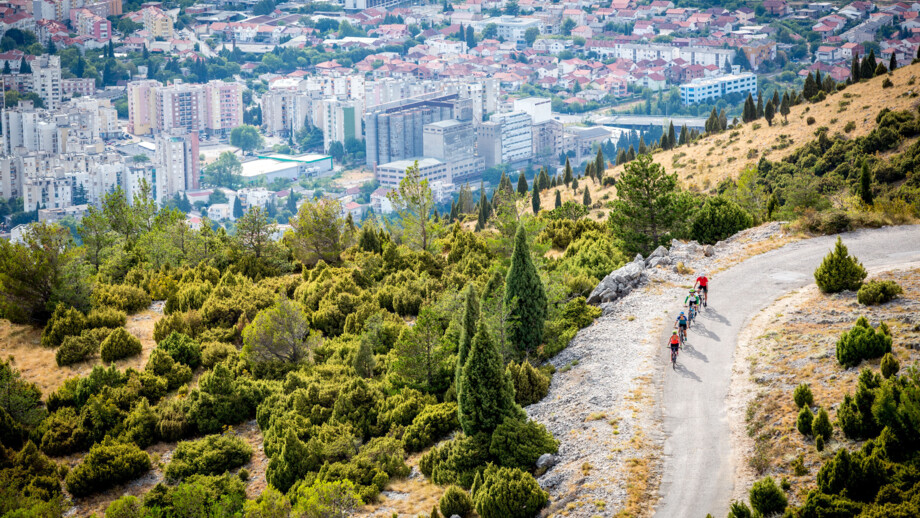 Drei Mountainbiker unterwegs durch Bosnien-Herzegowina. Im Hintergrund sieht man eine Stadt. Die drei Biker radeln auf einer Straße den Berg auswärts.