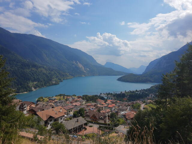 Ausblick auf den Molvenosee und Molveno. Die Sonne scheint. Der Himmel ist leicht bewölkt. Um den See erheben sich die Berge.