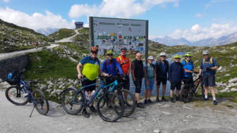 Eine Gruppe von Mountainbiker*innen am Pfitscherjoch. Sie stehen vor dem Schild umliegend ist die Bergwelt.