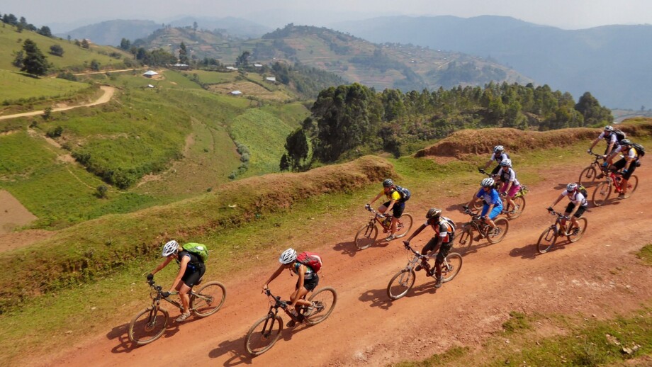 Eine Mountaibike-Gruppe fährt auf einer Sandpist in Höhenlage durch Ruanda. Im Hintergrund sieht man die Hügellandschaft.