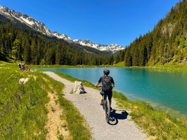 Mountainbiker am Herzsee im Kleinwalsertal. Der Himmel ist strahlend blau. Keine Bewölkung. Die Sonne scheint. Im Hintergrund sind die Gipfel der Allgäuer Bergwelt zu sehen.