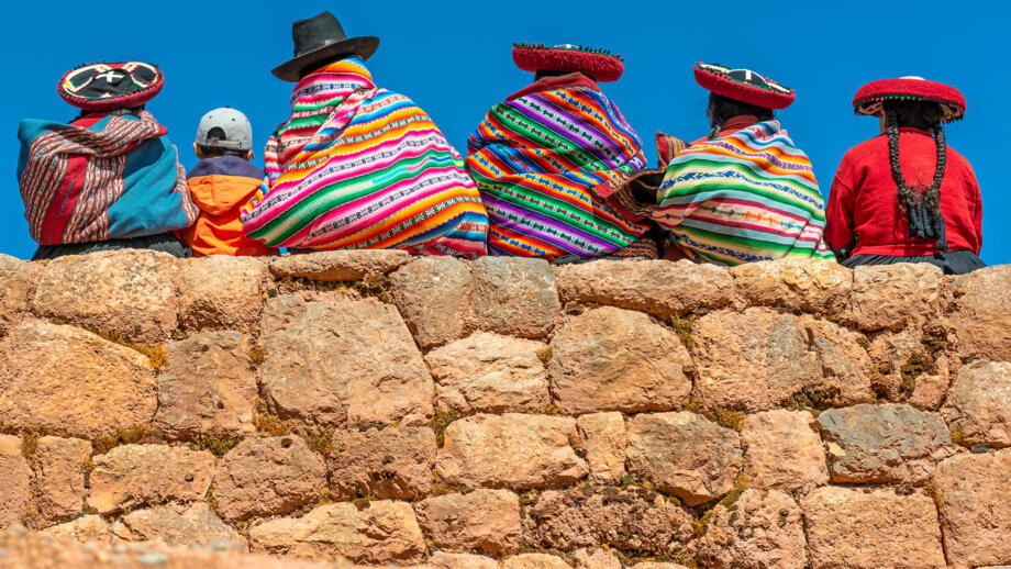 Vier einheimische mit traditionell-bunter Kleidung und mit Hüten auf einer alten Inkamauer sitzend, Aufnahme von Hinten