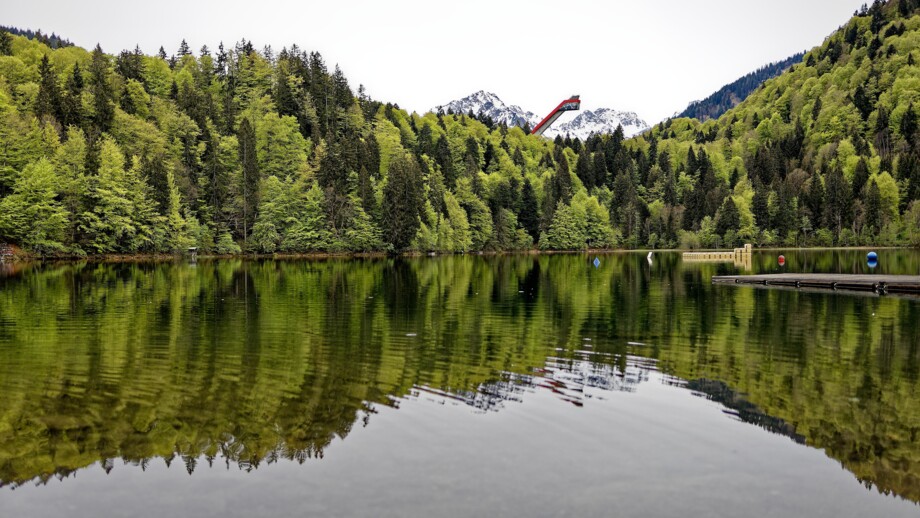 Ein ruhiger Bergsee spiegelt dichte grüne Wälder und schneebedeckte Gipfel wider, während im Hintergrund eine markante Skisprungschanze aus den Bäumen hervorragt.
