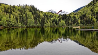 Ein ruhiger Bergsee spiegelt dichte grüne Wälder und schneebedeckte Gipfel wider, während im Hintergrund eine markante Skisprungschanze aus den Bäumen hervorragt.