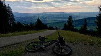 Ein Fahrrad liegt auf einem Bergweg mit weitem Blick über eine hügelige Landschaft, während Sonnenstrahlen dramatisch durch die Wolken am Abendhimmel brechen.