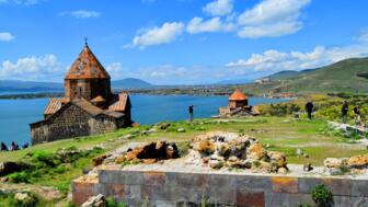 Zwei Kreuzkjuppelkirchen auf grünem Wiesengrund am tiefblauen Sevansee in Armenien