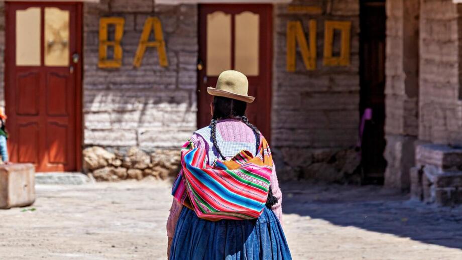 Bolivianier in traditionell bunter Tracht mit Hut, die Einkäufe in ihre einfache Behausung trägt