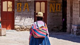 Bolivianier in traditionell bunter Tracht mit Hut, die Einkäufe in ihre einfache Behausung trägt