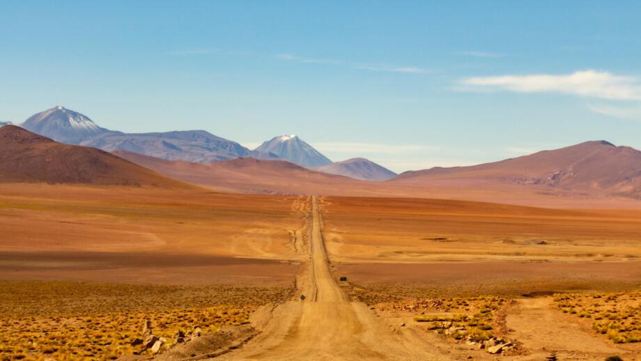 Wüstenpist auf rotem Sand in der Atacama mit Vulkanbergen im Hintergrund und blauem Himmel