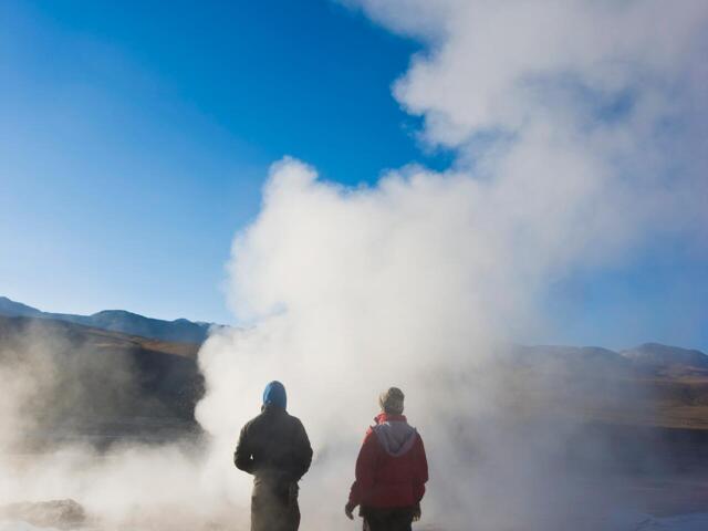Zwei Besucher von hinten blick auf einer der gischtenden Geysire des Vulkanfeldes El Tatio am Rand der chilenischen Atacama-Wüste