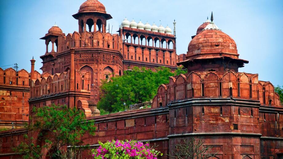 Das Rote Fort in Delhi ist eine weitläufige historische Festungsanlage aus rotem Sandstein mit hohen Mauern, Türmen und Kuppelbauten, die von grünen Bäumen und blühenden Pflanzen umgeben ist und sich vor einem klaren blauen Himmel erhebt.