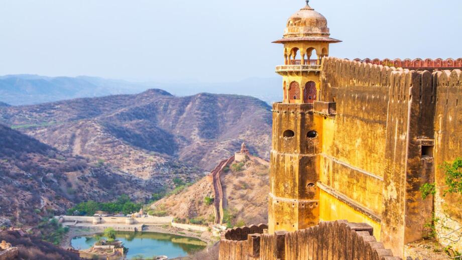 Amber Fort bei Jaipur ist eine imposante historische Festungsanlage auf einem Hügel, deren hohe sandfarbene Mauern sich entlang der kargen Berglandschaft erstrecken und einen Blick auf einen kleinen See im Tal freigeben.
