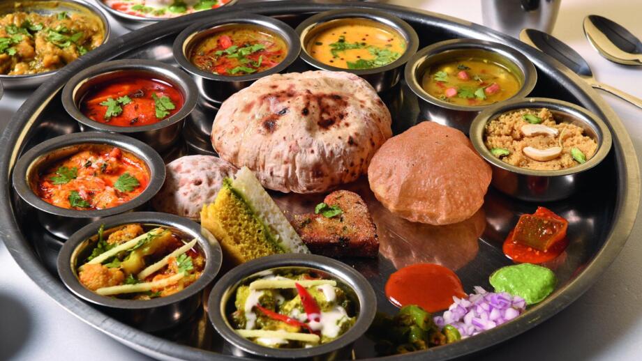 Zu sehen ist eine traditionelle indische Thali-Mahlzeit auf einer großen Metallplatte mit zahlreichen kleinen Schalen, gefüllt mit verschiedenen Currys, Gemüsegerichten, Chutneys, Brot und Süßspeisen.