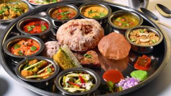 Zu sehen ist eine traditionelle indische Thali-Mahlzeit auf einer großen Metallplatte mit zahlreichen kleinen Schalen, gefüllt mit verschiedenen Currys, Gemüsegerichten, Chutneys, Brot und Süßspeisen.