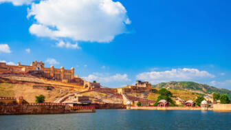 Zu sehen ist das Amber Fort bei Jaipur, eine weitläufige Festungsanlage aus hellem Sandstein, die sich über die Hügel Rajasthans erstreckt und malerisch an einem ruhigen See vor einem strahlend blauen Himmel liegt.