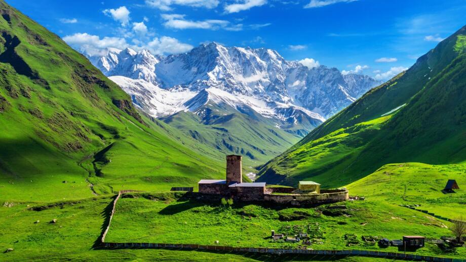 Bei Uschguli in Georgiens Provinz Swanetien mit grünen Feldern, Gehöft mit Turm und Steinmauern als Umfassung und dem vergletschen Berg Schchara im Hintergrund vor blauem Himmel
