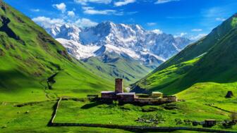 Bei Uschguli in Georgiens Provinz Swanetien mit grünen Feldern, Gehöft mit Turm und Steinmauern als Umfassung und dem vergletschen Berg Schchara im Hintergrund vor blauem Himmel