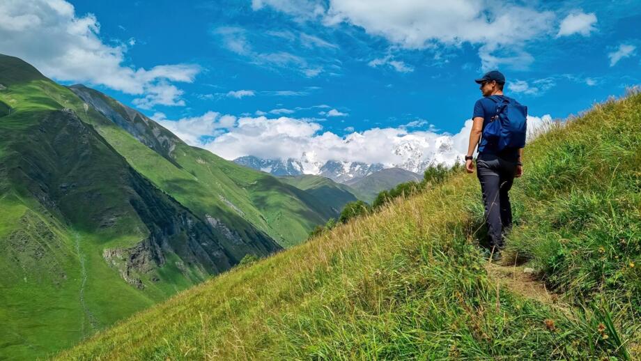 Wanderer mit blauem Shirt und Rucksack passiert eine grasbewachsenen Berghang nahe Uschguli in Swanetien und blick auf vergeltscherte Kaukasu-Gipfel