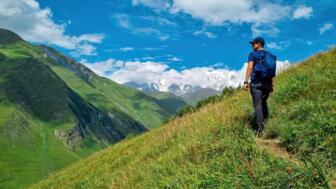 Wanderer mit blauem Shirt und Rucksack passiert eine grasbewachsenen Berghang nahe Uschguli in Swanetien und blick auf vergeltscherte Kaukasu-Gipfel