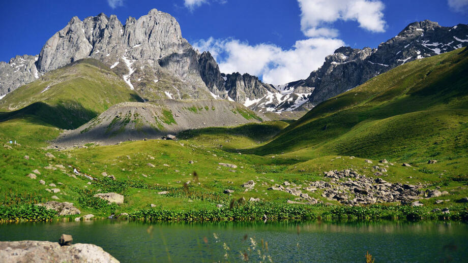 Georgisches Dorf Stepantsminda in grüner Wiesenlandschaft mit See im Vordergrund und felsigen Bergen im Hintergrund